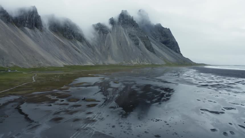Path winds through field toward mountains reflected in nearby wet beach sand on an overcast day