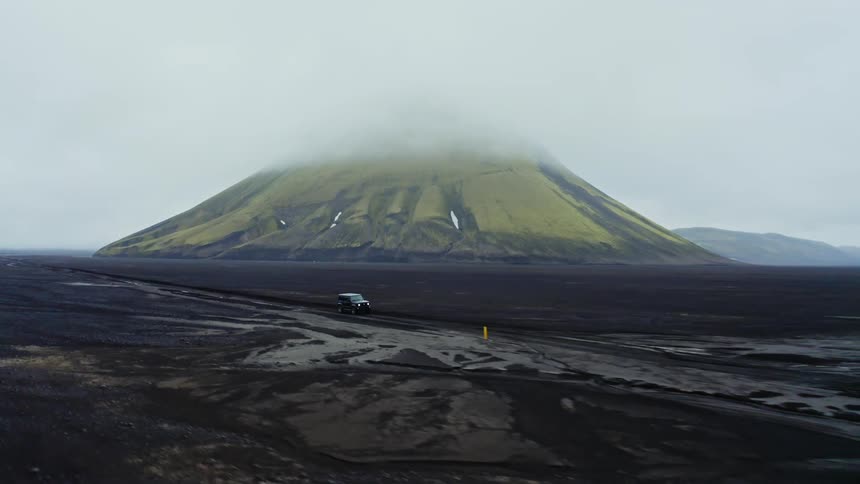 Jeep driving across icelandic terrain with green and black mountain in the background under a clouded sky