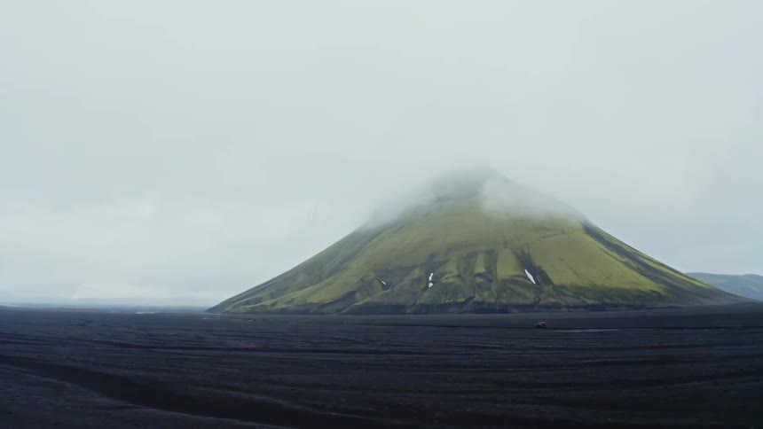 Vehicle drives along dark brown plain passing volcanic mountain under low lying clouds