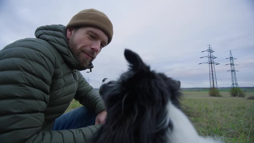 A man is bending down and playing with his dog in a rural grassland