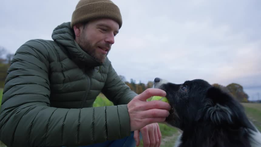 A bearded man wearing a brown hat and a green coat plays ball with his dog in a green field enjoying a simple and joyful moment of companionship