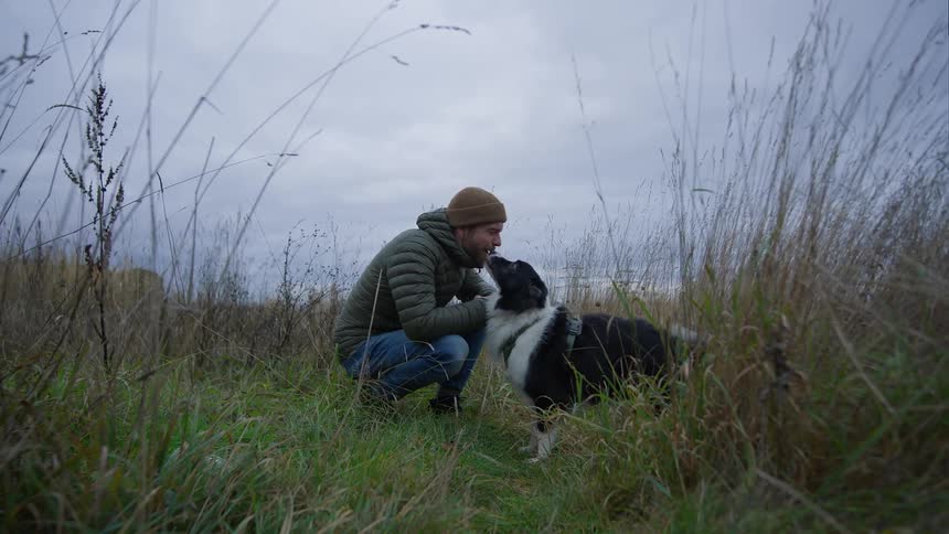 A man bends and plays with his dog in a rural grassland on a cloudy day
