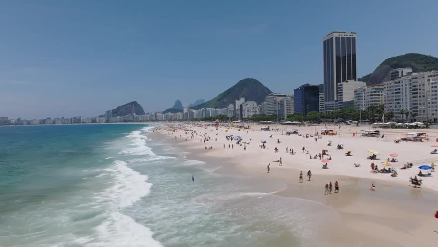 Waves crashing on a tourist filled beach lined with hotels on a sunny day under a blue sky with hills in the distance