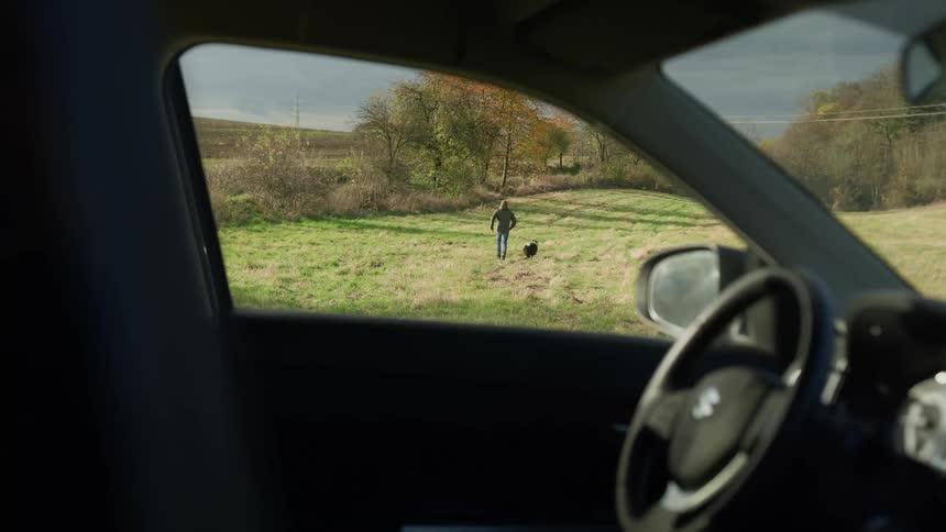 From his car a man plays with his dog in a grassy landscape