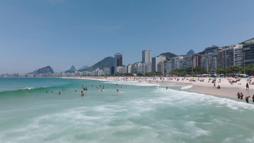 Waves roll onto crowded beach near buildings backed by mountains