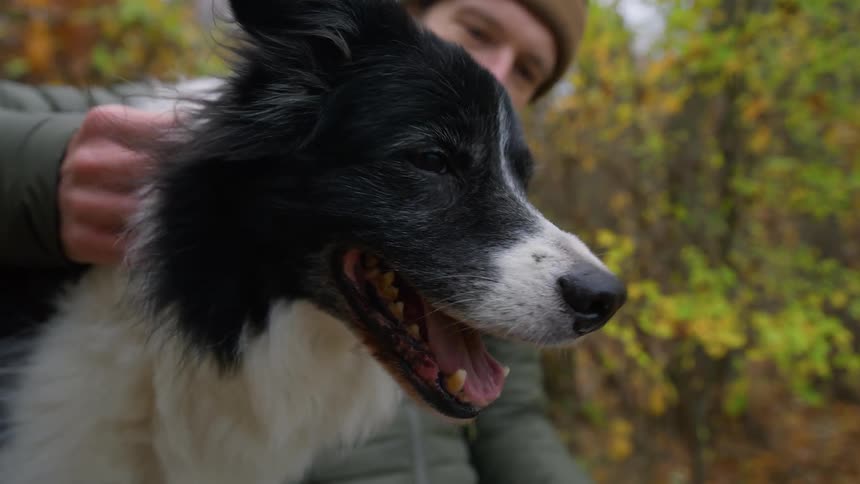 Man in a coat and beanie seated outdoors affectionately petting a dog by his side