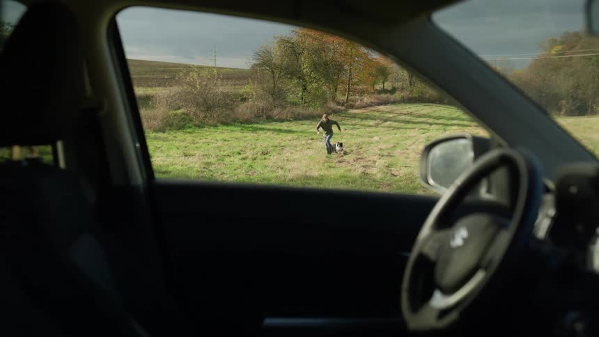 Man playing with a dog on a field during day time seen from the inside of a vehicle