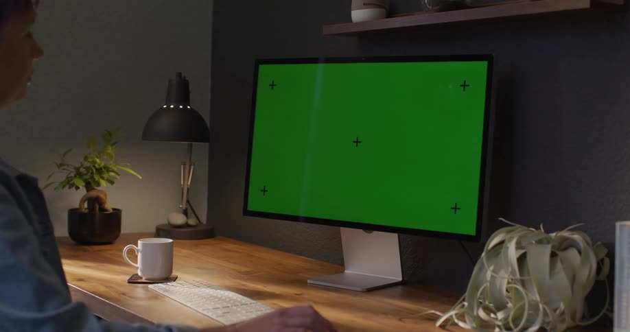 A woman is sitting at a table looking at a computer with a green screen