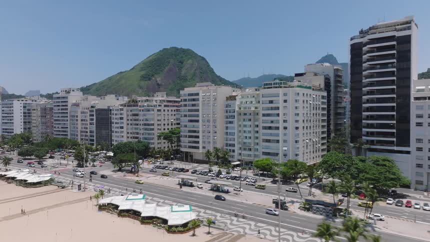 Traffic drives along street between beach and residential buildings in city nestled against mountains