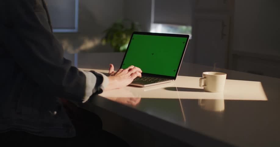 A person is sitting at a table typing on a laptop computer with a green screen as steam rises from a cup of coffee