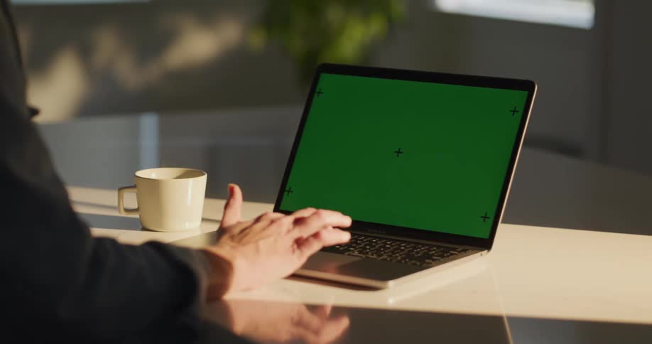 A woman is sitting at a counter yping on a laptop computer with a green screen as steam rises from a cup of coffee