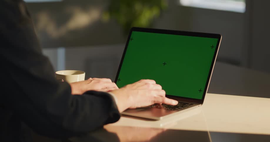 A man is sitting at a table typing on a laptop computer with a green screen
