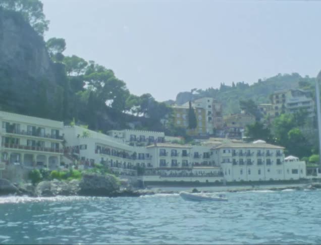 Woman with white hat and shirt travels on sailboat next to coastal town with buildings by cliff