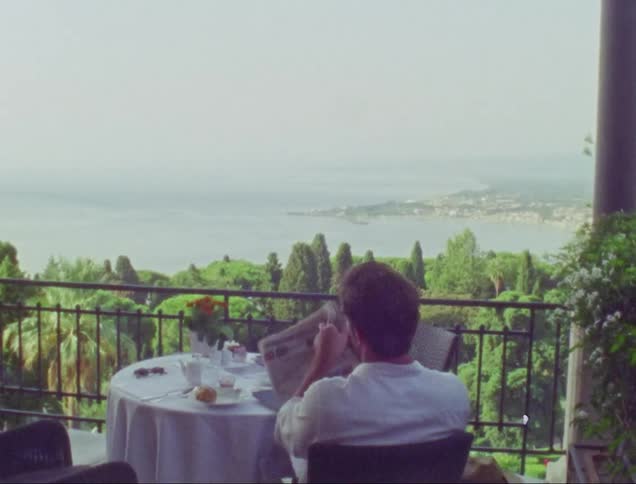 Man sits on terrace overlooking town and shore while reading the newspaper