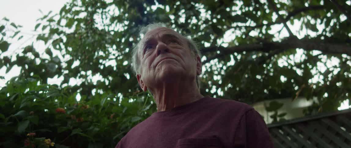 Elderly person sitting in an outdoor setting taking off their cap with emotion