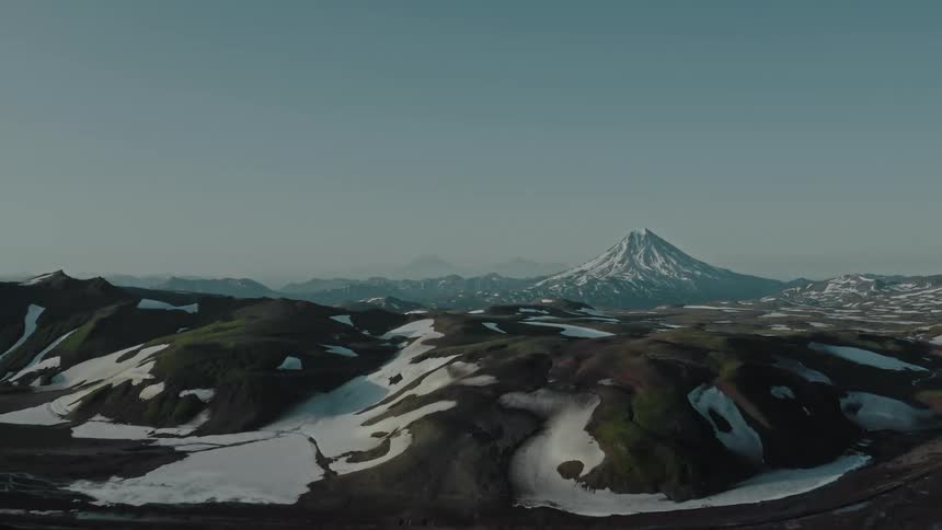 A tall mountain with snowy slopes behind a dark smooth rocky landscape with snow under an ombre blue sky during the day