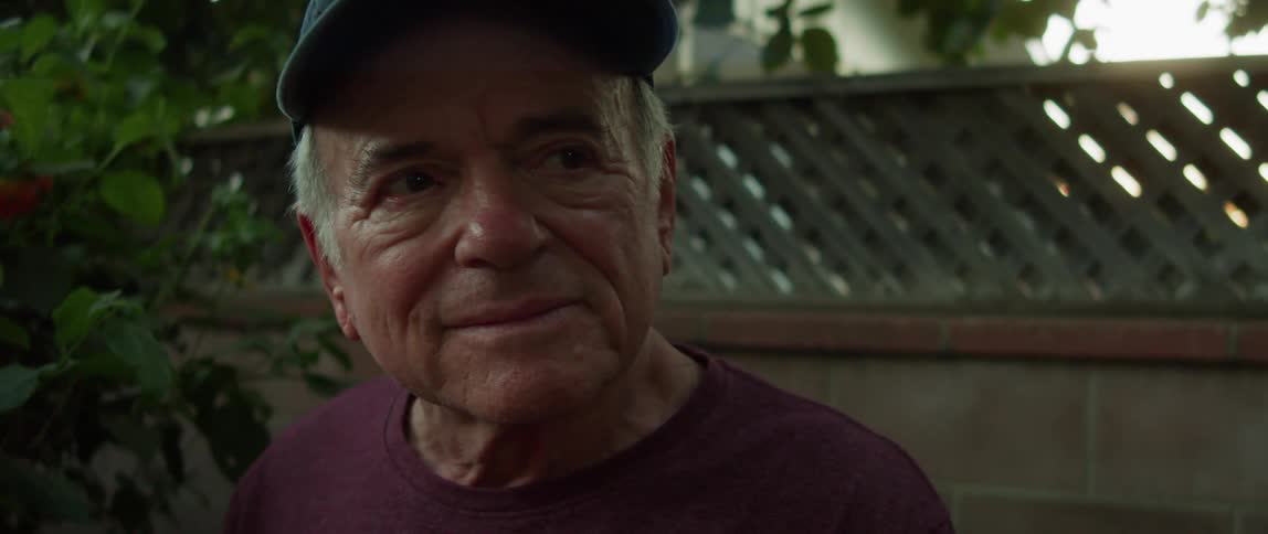 An elderly man is standing in a back yard looking intently to his left then looks to his right