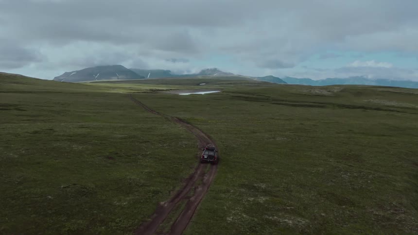 Aerial view of a jeep driving on a road in the hills next to the mountains