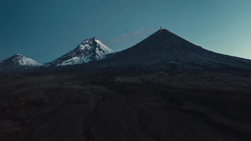 Panorama of a volcano erupting next to mountains and peaks covered in snow on a clear and blue sky day
