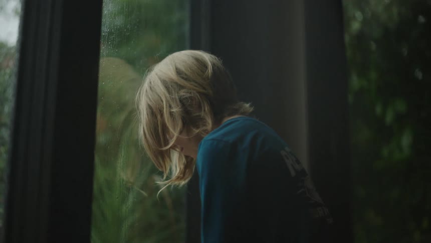 Sad blonde boy reclines on a window on a rainy day