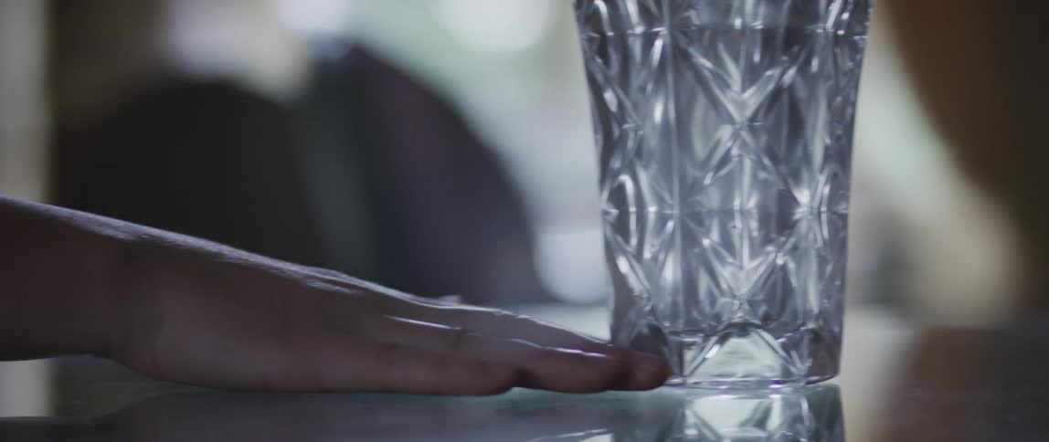 A hand pushing a glass vase across a table with the tip of its fingers