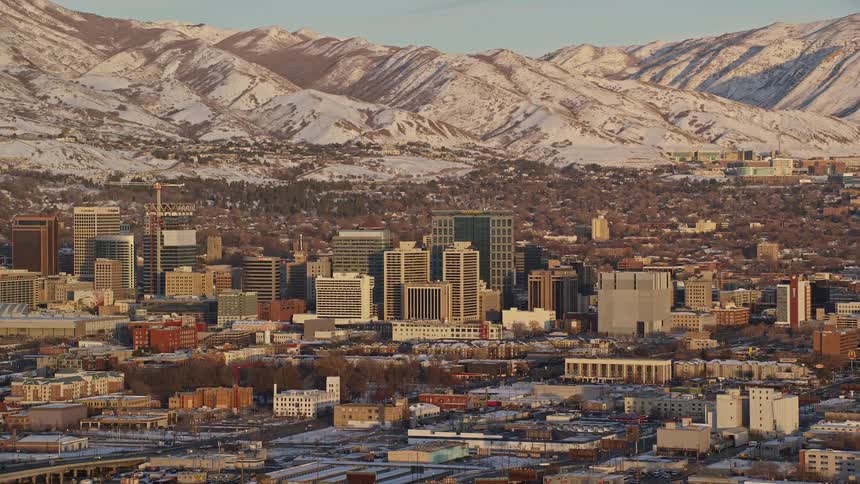 Aerial of neutral colored downtown skyline and city in front of snow covered neutral colored mountains on a bright day