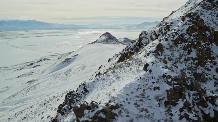 Aerial around the top of a snow and jagged brown rock covered mountain peak on a bright day with white snow covered land and a white clouded sky surrounding the background