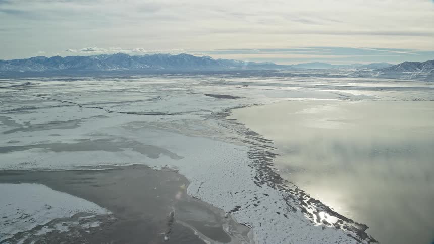 Aerial panorama of a snowcovered ground by the edge of a lake with a chain of mountains in the horizon and a morning sky