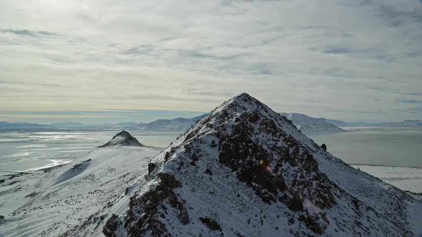 Aerial panorama of peaks and cliffs of a mountain range covered in snow by the edge of a lake in daylight
