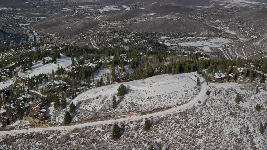 Aerial of large homes built between clusters of pine trees on a snowy mountain top with a valley behind it during the day