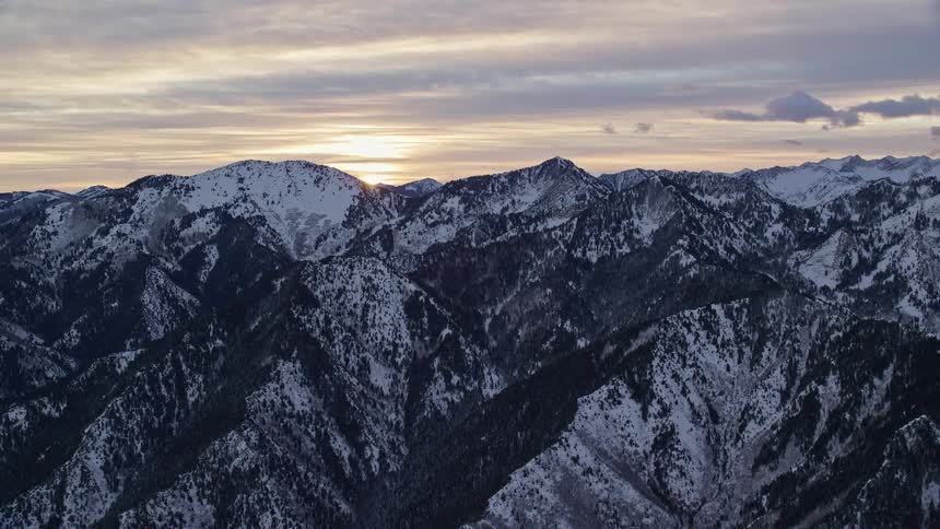 Snow covered mountains with mountains in the distance as the sun shines through the clouds on the horizon