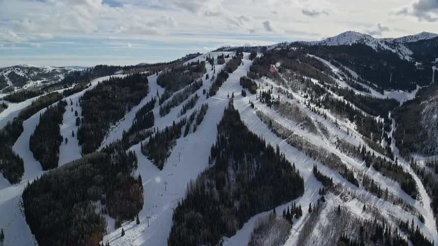 Aerial of a mountain with a ski lift up to the peak and multiple ski slopes covered in snow in the sunlight under a clouded blue sky