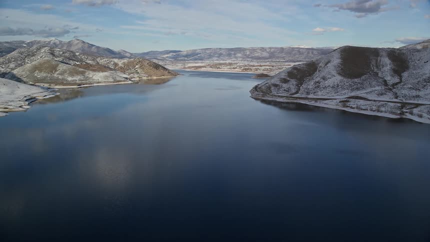Aerial over blue lake with clouds reflecting on it surrounded by snowy mountains under a bright blue sky