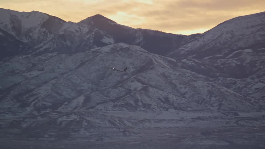 Airplane taking off against a backdrop of mountains partially covered in snow during sunrise