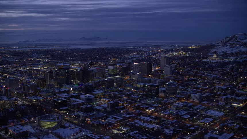 Aerial of a city expanding around tall centralized reaching a snow dusted mountain with the ocean on the horizon against a matching dark purpleblue sky at night