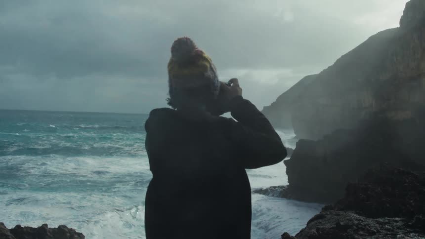 A person with a camera taking pictures of the view ahead the waves crashing into the rocks on a cloudy and windy day
