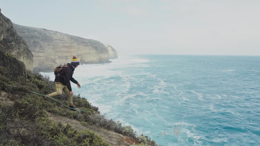A young male throws a climbing rope down the side of a cliff above the ocean