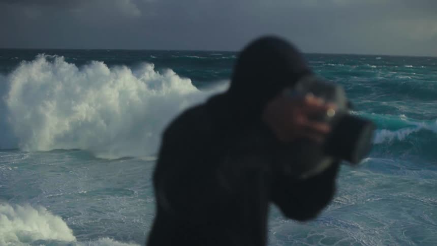 Man using a professional camera next to a turquoise blue sea with strong waves and wind