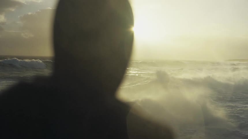 Close up ofthe back of a mans head as he watching the oceans rhythmic waves and the sun setting on the horizon