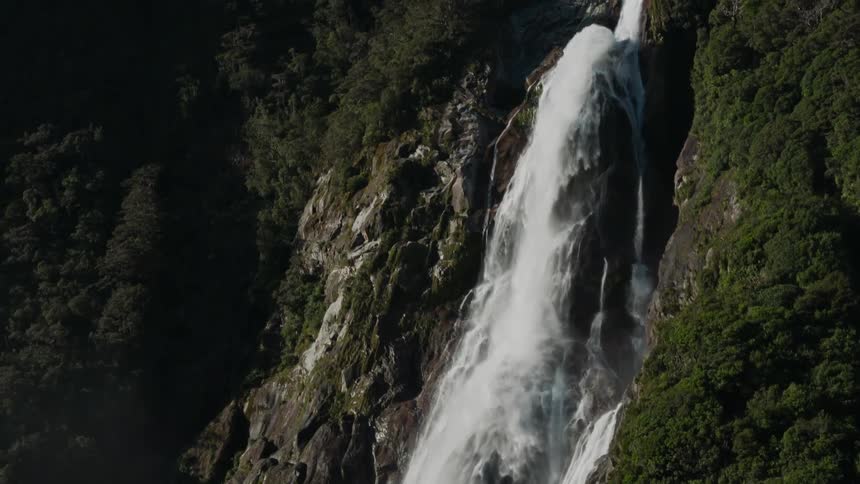 Waterfall surrounded by lush green vegetation up the side of the cliff to the top where a mountain peak sticks up above the land in the background under a blue sky on a sunny day
