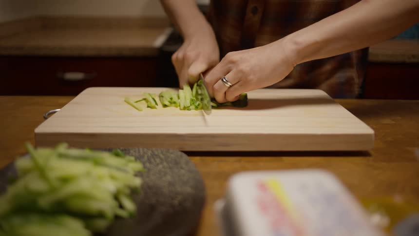 A person cutting cucumber into strips with a knife and cutting board in a kitchen