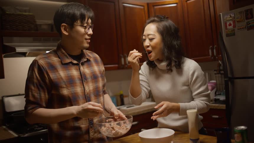 Two people in a kitchen are talking and tasting food in a pan