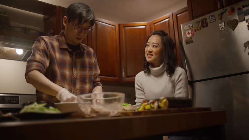 A man prepares a meal while talking to a woman in the kitchen