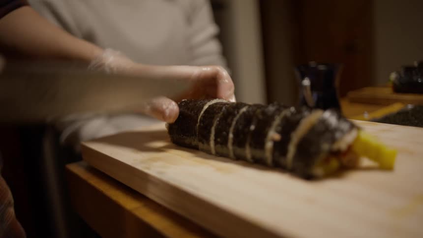 A persons hands slicing a roll of sushi on a wooden cutting board with a chefs knife