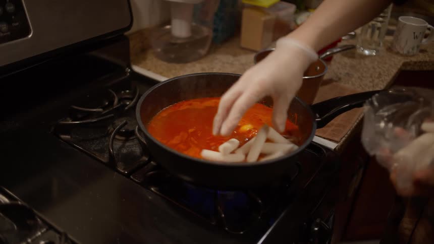 Closeup of a persons hands putting rice cakes into sauce in a pot on the stove