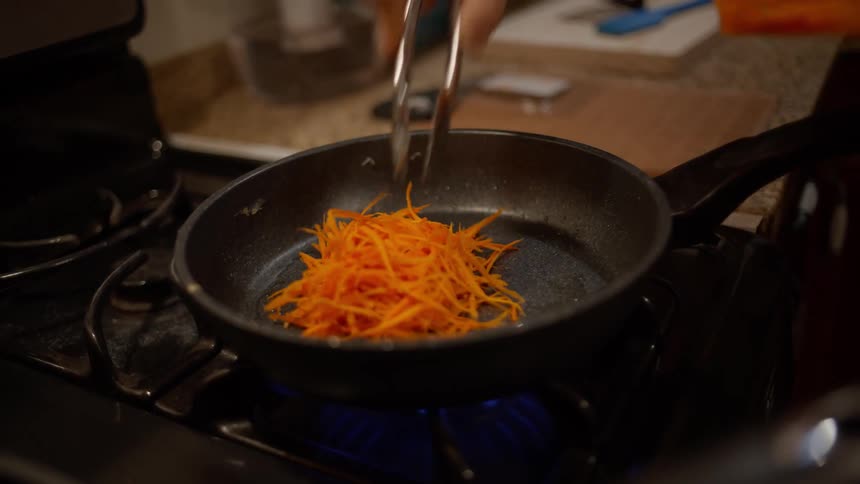 A hand using tongs to put sliced carrot into a frying pan on a stove
