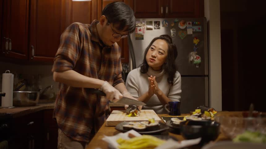 Woman sitting at a kitchen island counter laughing and talking with a man standing at the counter beside her cutting a sushi roll
