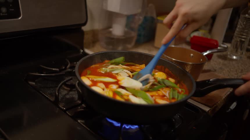 A persons hand stirring vegetables in a saucepan over a gas stove with a blue spatula