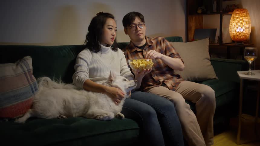 An asian couple sitting on their couch along with their dog while watching late night movie and eating popcorn