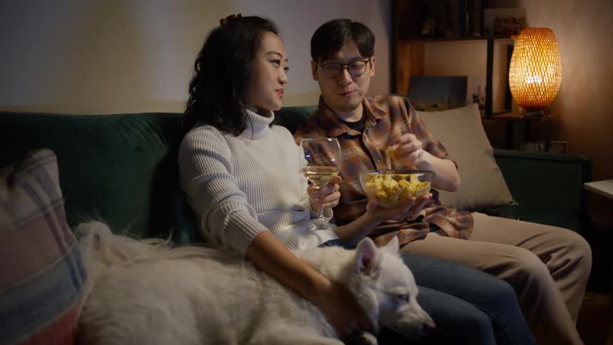 Asian man and woman sitting on the couch with their dog having fun enjoying snacks and drinks while watching tv